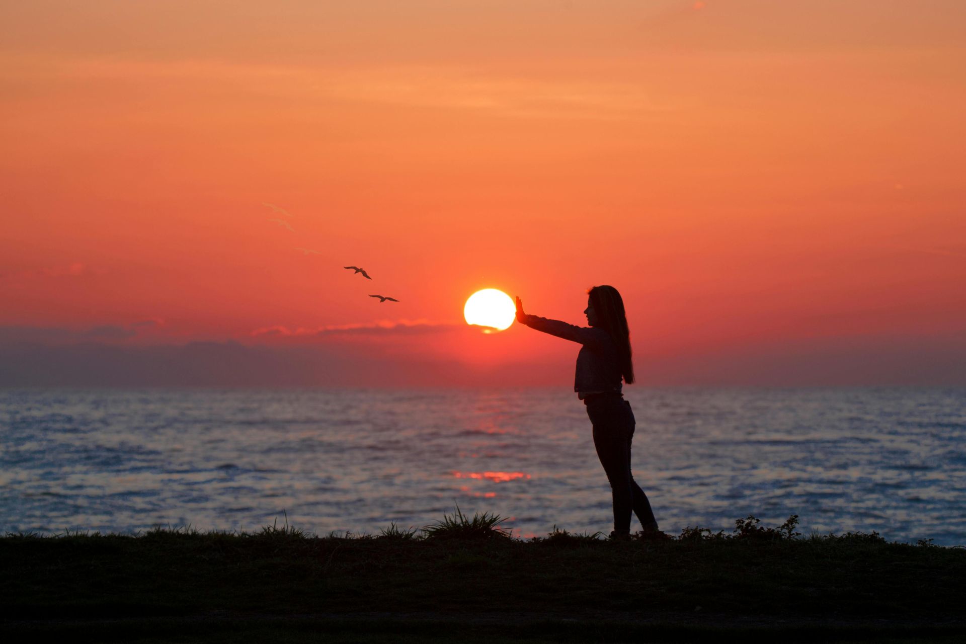 A woman silhouetted by the sunset, standing by the ocean, creating a tranquil seaside scene.