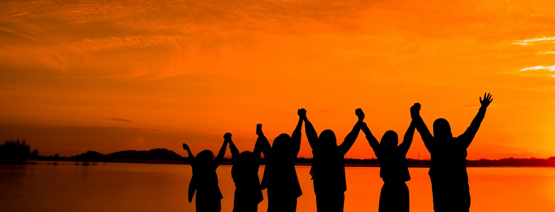 Silhouette of a group of friends with raised hands celebrating together by the lake during a beautiful sunset.